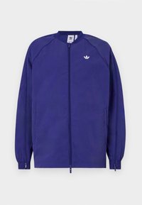 Lila lättviktig jacka med dragkedja framtill, ribbad krage och elastiska ärmslut. Har en liten vit logotyp på vänster bröst.