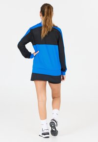 Blå och svart idrottsjacka med långa ärmar och hög krage. Slät textur, kontrasterande färger och en figurnära design.