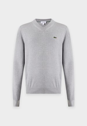 Joven con suéter de cuello en V gris claro sobre camisa blanca, vaqueros azules, cinturón negro y zapatillas blancas, de pie con una mano en el bolsillo.