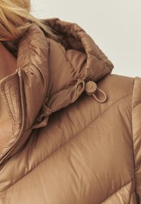 Doudoune marron avec un design matelassé, dotée d'une capuche à cordon de serrage et d'un bouton toggle circulaire. Texture douce et lisse avec des coutures visibles.
