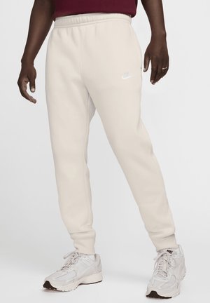 Light beige Nike sweatpants with an elastic waistband, featuring a white logo on the left thigh. Complemented by white sneakers.