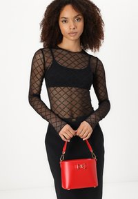 Blusa preta de malha de mangas longas com um padrão de diamante, combinada com uma saia justa preta. Segurando uma pequena bolsa de couro vermelha com detalhes em dourado.