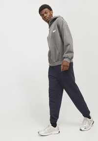 Grå bomulds hoodie med en frontlomme og broderet logo, kombineret med marineblå sweatpants. Modellen bærer sølv sneakers med sorte accenter.