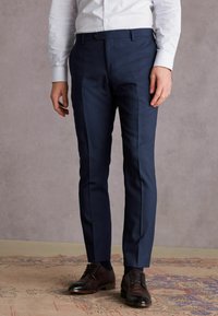 Navy-Hosen mit maßgeschneiderter Passform, die eine glatte Textur und einen dezenten Knick auf der Vorderseite aufweisen. Kombiniert mit dunkelbraunen Lederschuhen.