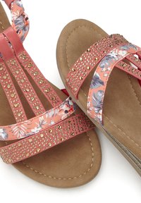 Koraalrode sandalen met bloemenstof banden, goudkleurige studs en een zachte bruine binnenzool met stikseldetails. Plat ontwerp voor comfort.