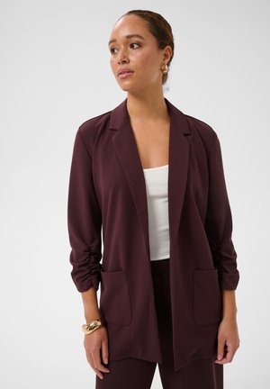 Kvinde iført en bordeaux blazer og bukser med en hvid top, guldfarvede øreringe og et kraftigt guldfarvet armbånd, der kigger til siden.