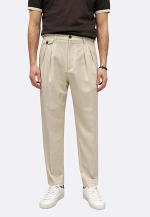 Uomo che indossa pantaloni beige plissettati, scarpe da ginnastica bianche e una camicia nera a maniche corte, in piedi davanti a uno sfondo neutro.