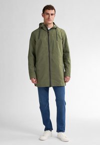 Groene waterdichte jas met een capuchon, trekkoorden en een voorkleinie. Gepaard met blauwe spijkerbroeken en witte sneakers. Soepele textuur.