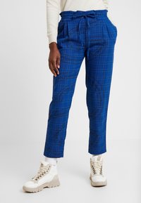 Blå rutig byxor har dragsko i midjan, sidofickor och svarta textmönster. Kombineras med beige och vita sneakers.