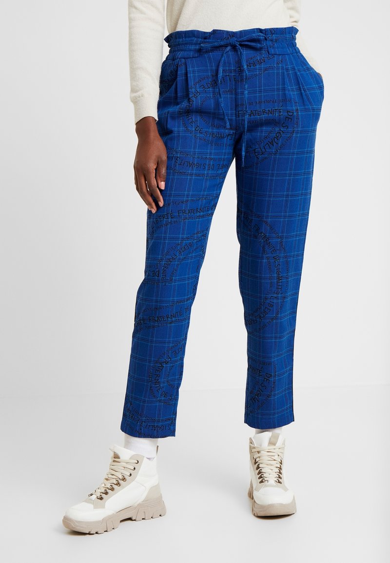 Blå rutig byxor har dragsko i midjan, sidofickor och svarta textmönster. Kombineras med beige och vita sneakers.