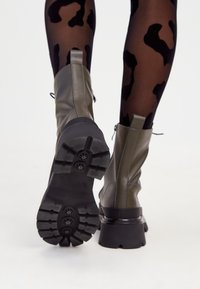 Bottines en cuir vert olive avec une semelle noire épaisse et une texture antidérapante. Le design comprend une fermeture éclair sur le côté et une toe arrondie.