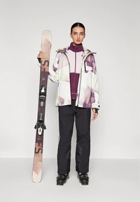 Snö sportutrustning med en mönstrad vit och lila jacka, ett lila baslager, svarta byxor och en skida med bindningar.