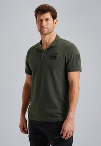 Polo de color verde oliva fabricado en algodón, con una tapeta de tres botones, mangas cortas y dos parches bordados en el pecho.