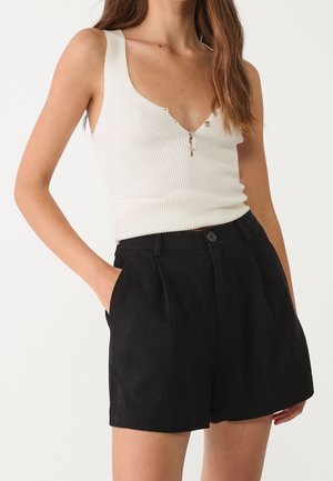 Vrouw in een mouwloze witte geribbelde top met knoopdetails, gecombineerd met hoge zwarte op maat gemaakte shorts, met handen in de zakken, tegen een witte achtergrond.