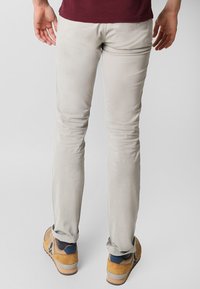 Lys beige slim-fit bukser lavet af bomuld, med en glat tekstur og subtile syninger, parret med brune og blå sneakers.