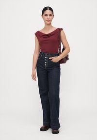 Burgunderfarget ermeløs topp med drapert halslinje, høyt liv mørk denim flared jeans med knappemulsjon foran, og runde svarte sko.