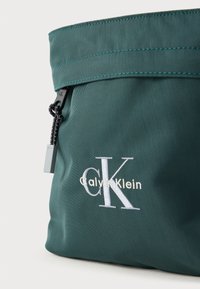 Zöld nejlontáska, átfedős kialakítással, hímzett "Calvin Klein" logóval és cipzáras záródással, texturált húzófüllel.