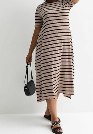 Vrouw draagt een midi-jurk met beige en zwarte horizontale strepen, houdt een kleine zwarte handtas vast en draagt zwarte sandalen met een gouden gesp.