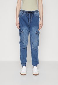 Person som bär blå denim cargo joggerbyxor med elastiska muddar, fram- och sidofickor, samt vita sneakers, stående mot en neutral bakgrund.