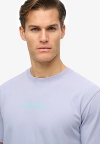 Camiseta de algodón de color morado claro con cuello redondo y un pequeño logo "Superdry" en color verde menta centrado en el pecho.