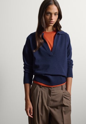 Jonge vrouw met lang bruin haar, gekleed in een marineblauwe V-hals trui over een oranje blouse en bruine geplooide broek tegen een effen achtergrond.