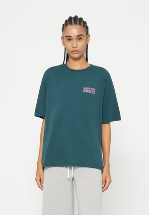 Jeune femme aux cheveux tressés portant un t-shirt oversize vert foncé avec un texte rose indiquant « "Forever chasing sunsets" » et un pantalon gris clair.