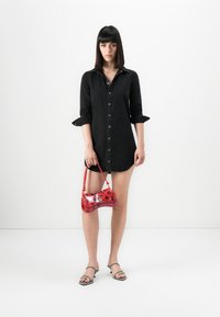 Woman in black buttoned shirt dress holding red rose-print handbag, wearing black strappy heeled sandals, standing against white background.