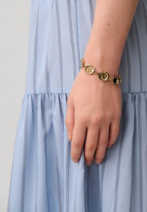 Main avec un bracelet en maillons dorés reposant contre un tissu de robe bleu clair à rayures.