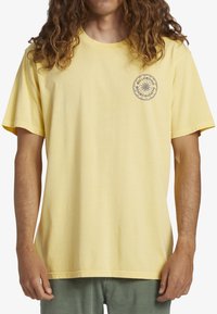 Camiseta de algodón amarilla con un ajuste relajado, mangas cortas y cuello redondo. Presenta un logotipo gráfico circular en el lado izquierdo del pecho.