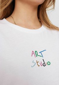 Biela bavlnená tričko s okrúhlym výstrihom, na ktorom je viacfarebný vyšívaný text "ART studio" v hravých písmach.