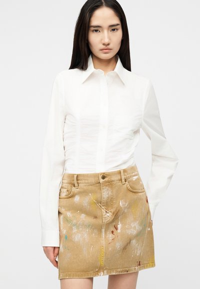 Γυναίκα που φοράει εφαρμοστό λευκό πουκάμισο με κουμπιά και μπεζ φούστα τζιν με πιτσιλιές από μπογιά, στέκεται μπροστά σε απλό φόντο.
