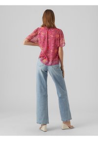 Blouse semi-transparent à motifs floraux roses avec manches courtes, associée à un jean taille haute bleu clair et des sandales blanches à talons carrés.