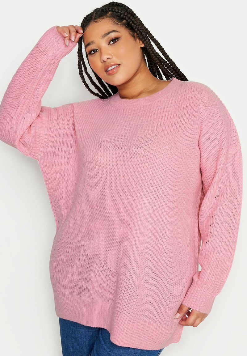 Överdimensionerad rosa stickad tröja med ett strukturerat mönster, ribbade ärmmuddar och rundad halsringning, kombinerad med mörkblå jeans.