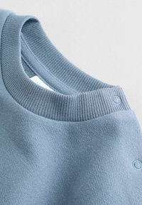 Błękitny sweatshirt z rypsowym dekoltem typu crew. Charakteryzuje się gładką fakturą i widocznymi szwami. Zawiera dwa zatrzaski w okolicy ramienia.