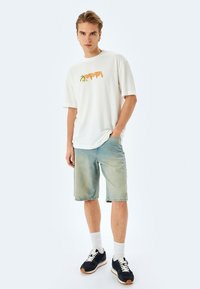 Hvid kortærmet t-shirt med orange og grøn grafisk design, parret med lyseblå denimshorts, hvide sokker og sorte sneakers.