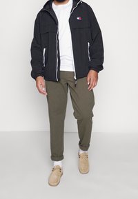 Lekka czarna kurtka z zamkiem z przodu, dwiema kieszeniami, elastycznymi mankietami, noszona na białej koszulce; w połączeniu z oliwkowozielonymi spodniami i beżowymi butami.