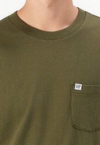 Közelkép a nyakról és a felső krékről, amelyen egy olíva zöld póló látható, jobb oldalon egy kis GAP felirattal ellátott zsebbe.