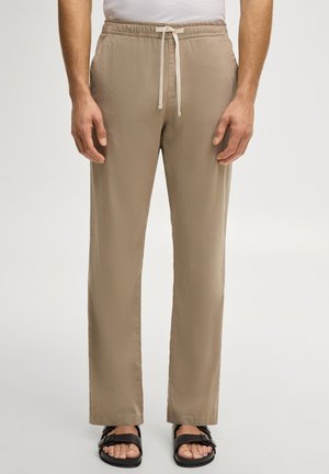 Man die beige broek met trekkoord en zwarte sandalen met gesp draagt, staand tegen een effen lichte achtergrond.