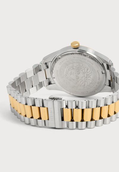 Bracelet de montre en métal argenté et doré avec fermoir marqué Versace, montrant le fond de montre en acier inoxydable gravé avec un motif de tête de Méduse.