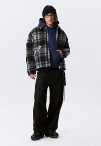 Jong persoon met een zwarte beanie, bril, marineblauwe hoodie, zwart-wit geruite jas, losse zwarte broek met ketting en zwarte schoenen die tegen een witte achtergrond staat.