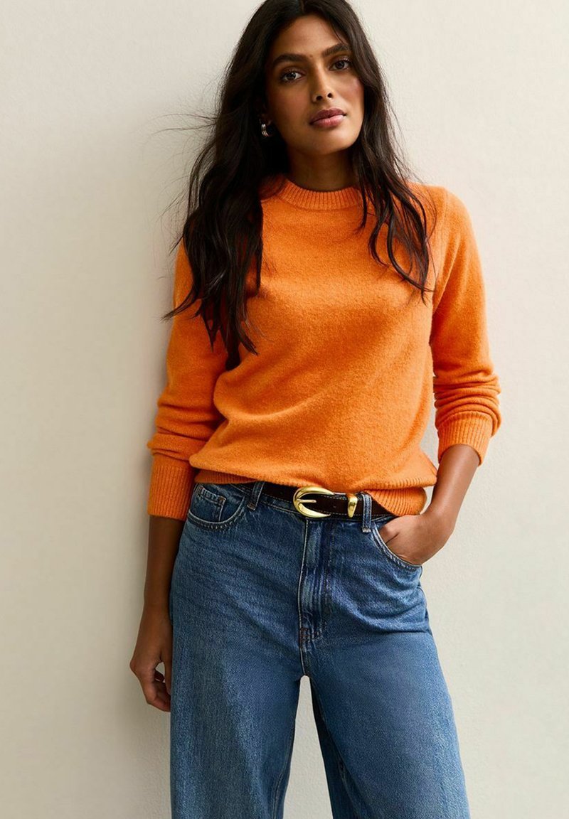 Femme aux longs cheveux bruns, portant un pull orange et un jean bleu avec une ceinture noire, se tenant devant un mur clair et uni.