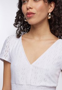 Abito bianco con scollatura a V, maniche corte a puff e un motivo a occhio testurizzato. Grandi orecchini a cerchio in oro completano il look.
