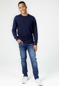 Granatowy sweter z okrągłym dekoltem, ze ściągaczami, noszony na jasnoniebieskich dżinsach. Model stoi swobodnie, z jedną ręką w kieszeni, w białych trampkach.