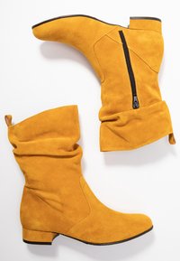 Bottines en suede jaunes avec un design à fronces, fermeture éclair latérale et petit talon bloc. Texture lisse et détails de couture soignés.