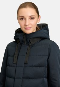 Marineblauwe pufferjack met een capuchon, doorgestikte ontwerpen en zwarte verstelbare koorden. Gladde textuur met een ritssluiting aan de voorkant.