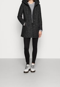 Cappotto grigio con texture, cappuccio, zip frontale e tasche. Indossato sopra un maglione leggero e leggings neri, abbinato a stivali bianchi alla caviglia.