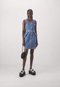 Denim mini kleita ar pieguļošu augšdaļu, kvadrātu izgriezumu, divām priekšējām kabatām un priekšējo pogu aizvēršanu. Apvienota ar masīvu melnu platformas sandali.