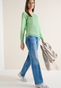 Femme marchant, vêtue d'un pull vert, d'un jean bleu et de baskets argentées, tenant une veste beige dans la main droite, sur un fond clair uni.