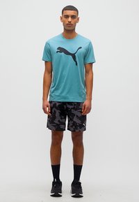 Νεαρός άνδρας στέκεται, φορώντας μπλε-πράσινη μπλούζα Puma, μαύρα σορτς με καμουφλάζ, μαύρες κάλτσες και μαύρα αθλητικά παπούτσια, μπροστά σε απλό φωτεινό φόντο.