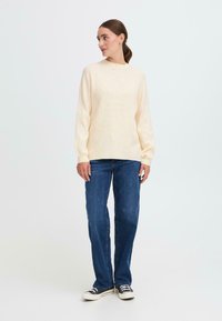 Cremfarbener Rippenpullover mit langen Ärmeln, kombiniert mit blauen geraden Jeans und schwarz-weißen gemusterten Sneakers.